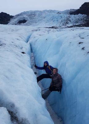Two happy girls are sitting inside a blue ice crevasse, with their feet on the walll.