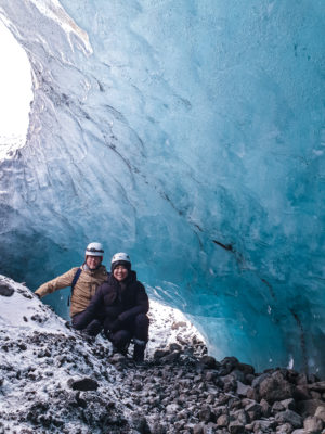 two people wearing glacier equipment kneeling down in a blue ice cave