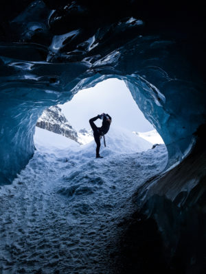 a girl wearing glacier gear doing a gymnastic pose at the entrance of a blue ice cave on a glacier