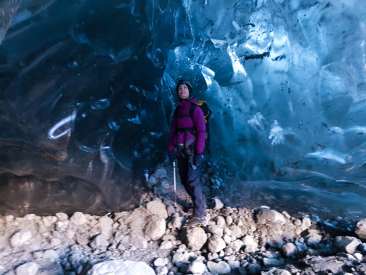 a girl wearing glacier equipment, crampons, harness, helmet, ice axe and a backpack stands beneath a blue ice wall in an ice cave